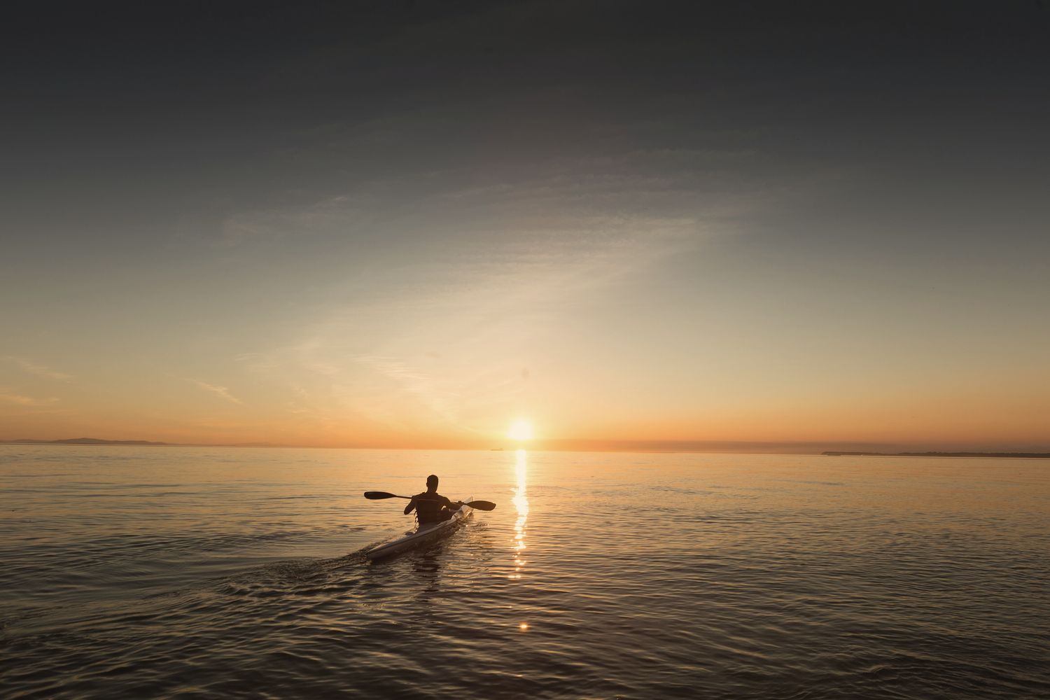 A kayaker heading out across a calm bay into the setting sun.